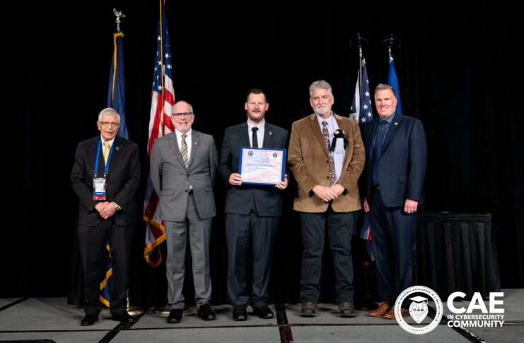Five men in suits stand on stage with flags, one holding a certificate; CAE in Cybersecurity Community logo is visible.