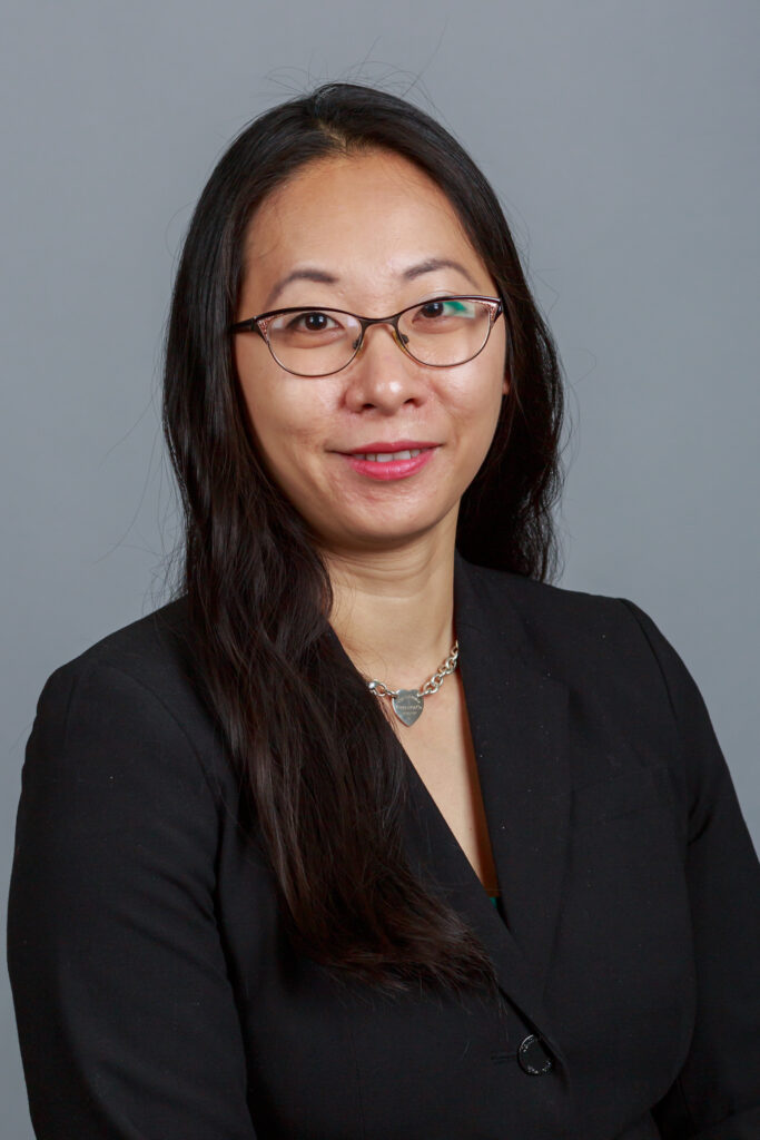 Woman with long black hair and glasses wearing a black blazer and necklace, posed against a gray background.