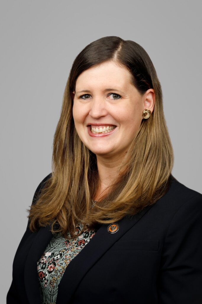 Woman with long brown hair, wearing a dark blazer, patterned top, and gold earrings, smiling at the camera.