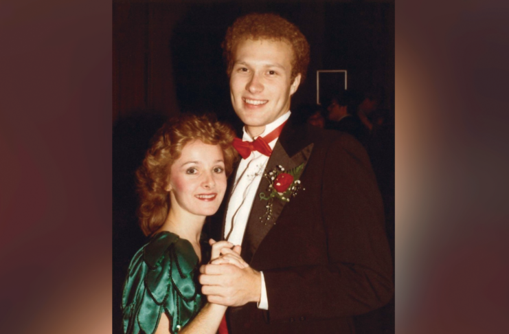A young couple in formal attire pose together while dancing, with the man wearing a tuxedo and the woman in a green dress.