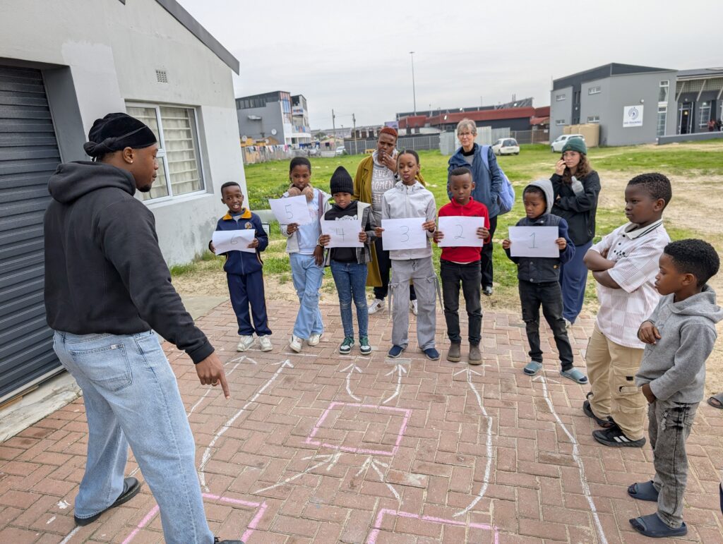 A group of children holding numbered sheets stand in line outside as a man instructs them near a chalk hopscotch grid.