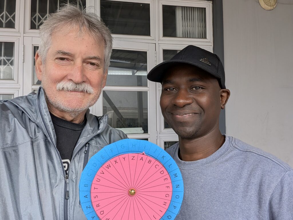 Two men standing together, holding a colorful alphabet spinner, smiling at the camera in front of a windowed wall.