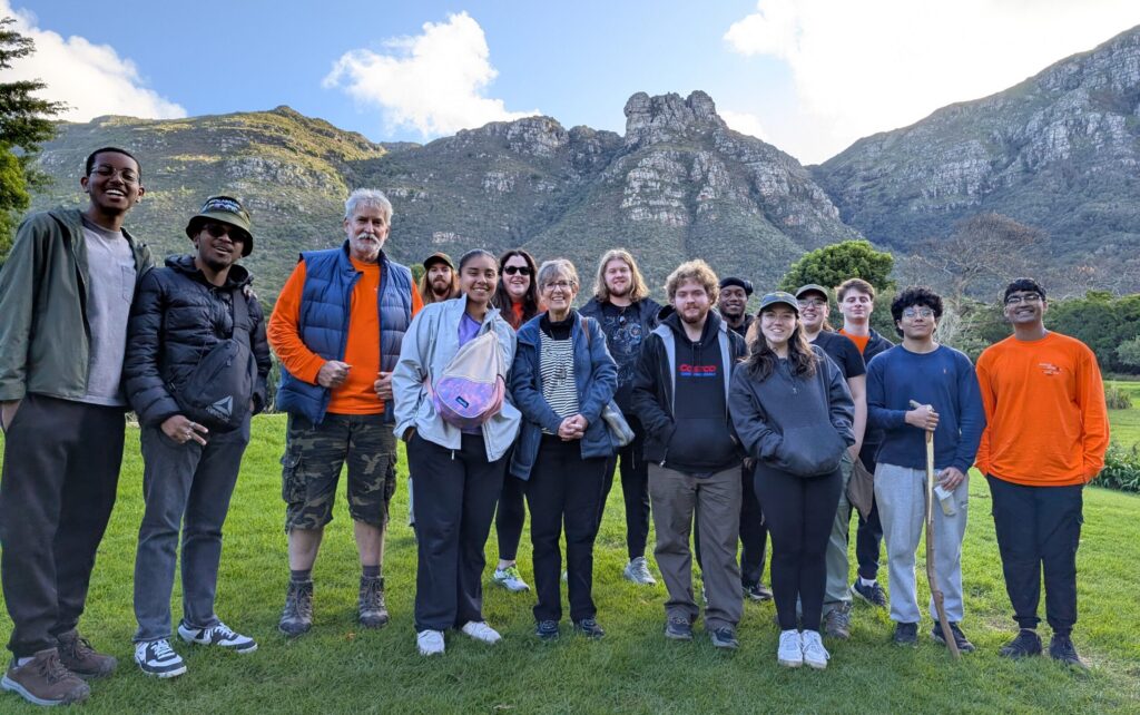 A group of people stand together on grass with mountains and blue sky in the background.