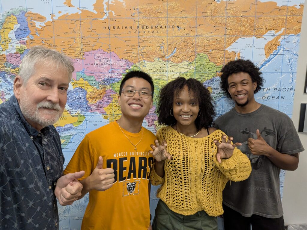 Four people standing in front of a world map, smiling and making hand gestures toward the camera.