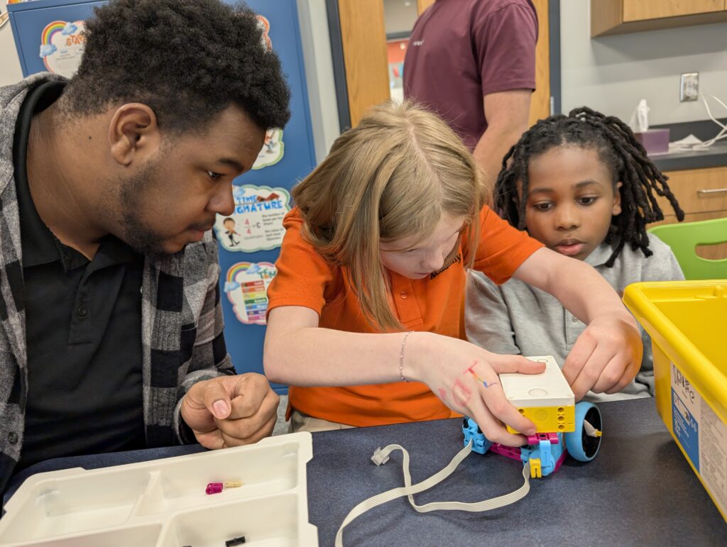 Three children work together assembling a small robot on a table in a classroom setting.