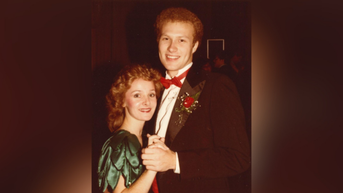 A young couple in formal attire pose together while dancing, with the man wearing a tuxedo and the woman in a green dress.