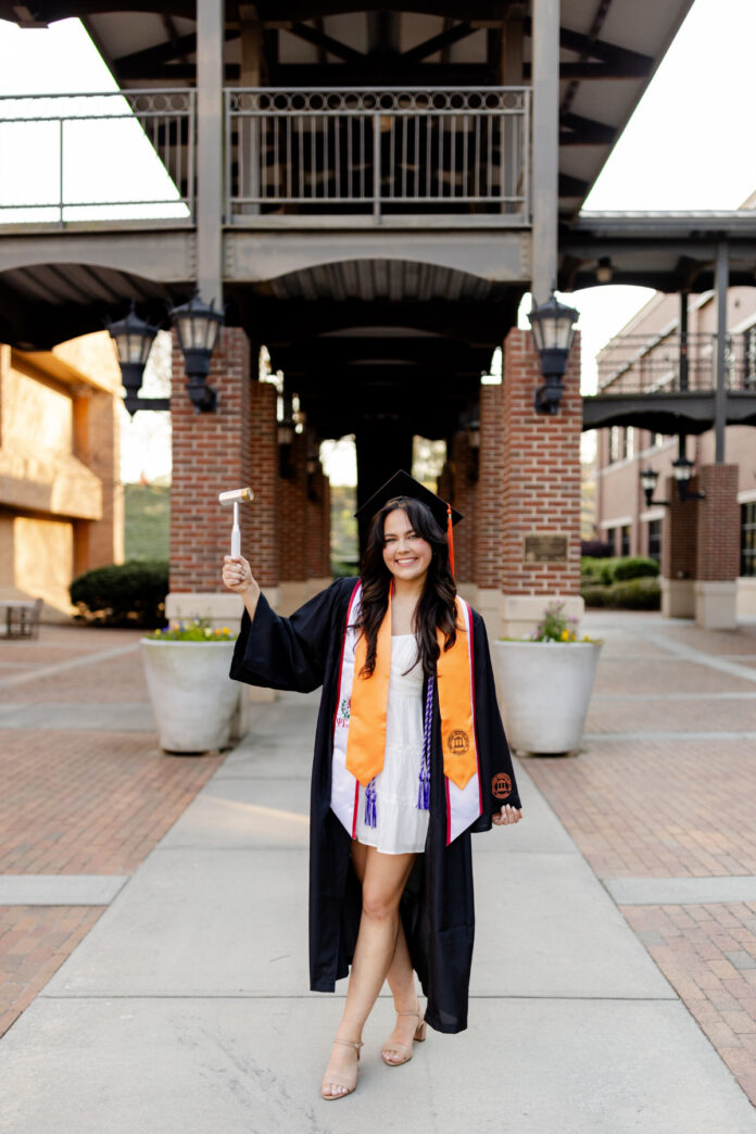 Samanta Schultz wears graduation attire and stands outdoors, holding a hammer and smiling at the camera.