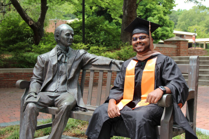 Samuel Pulapakuri, in cap and gown, sits smiling on a bench next to the Jesse Mercer statue in Atlanta.