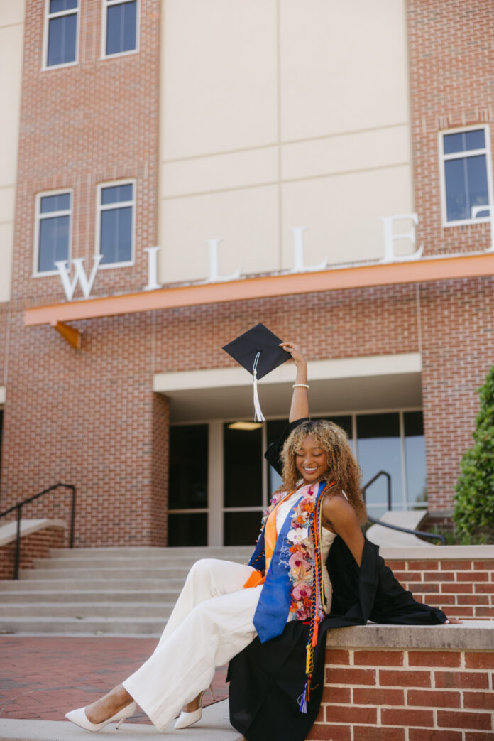 Teresita Cabral in cap and gown sits on steps outside the Willet Science Center, holding up her cap and smiling.