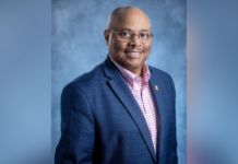 Music alumnus receives national recognition for library work A man in a navy blazer and pink checkered shirt poses against a blue-gray studio background, smiling at the camera.
