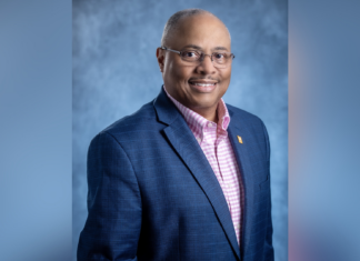 Music alumnus receives national recognition for library work A man in a navy blazer and pink checkered shirt poses against a blue-gray studio background, smiling at the camera.