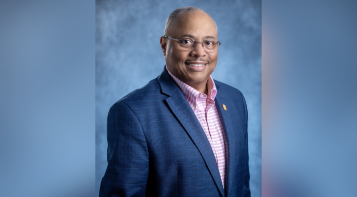 Music alumnus receives national recognition for library work A man in a navy blazer and pink checkered shirt poses against a blue-gray studio background, smiling at the camera.