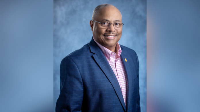 A man in a navy blazer and pink checkered shirt poses against a blue-gray studio background, smiling at the camera.
