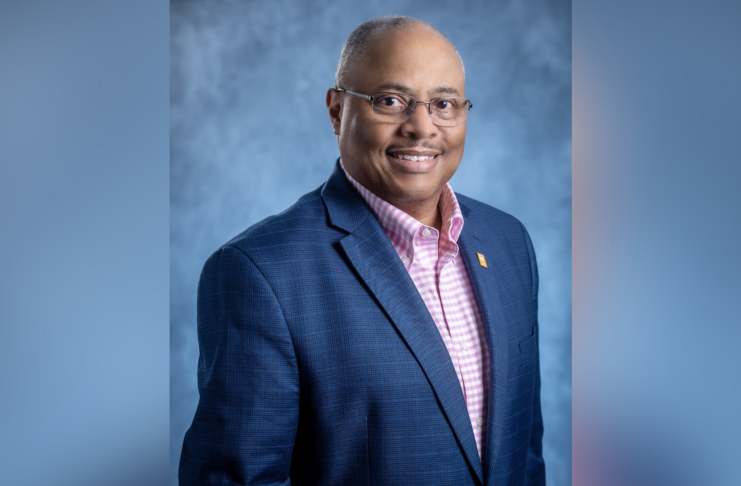 A man in a navy blazer and pink checkered shirt poses against a blue-gray studio background, smiling at the camera.