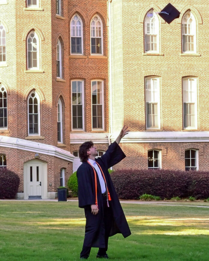 Graduate in cap and gown throws graduation cap into the air in front of a brick academic building.