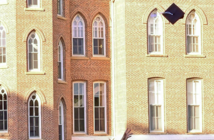 Graduate in cap and gown throws graduation cap into the air in front of a brick academic building.