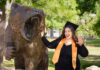 Graduate Spotlight: Cailey Martinez A graduate in a cap and gown poses next to a roaring bear statue outdoors, smiling and holding up one hand.