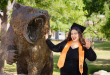 Graduate Spotlight: Cailey Martinez A graduate in a cap and gown poses next to a roaring bear statue outdoors, smiling and holding up one hand.