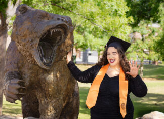 Graduate Spotlight: Cailey Martinez A graduate in a cap and gown poses next to a roaring bear statue outdoors, smiling and holding up one hand.