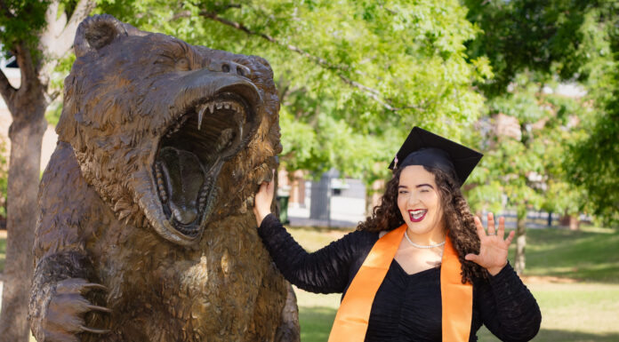 Graduate Spotlight: Cailey Martinez A graduate in a cap and gown poses next to a roaring bear statue outdoors, smiling and holding up one hand.