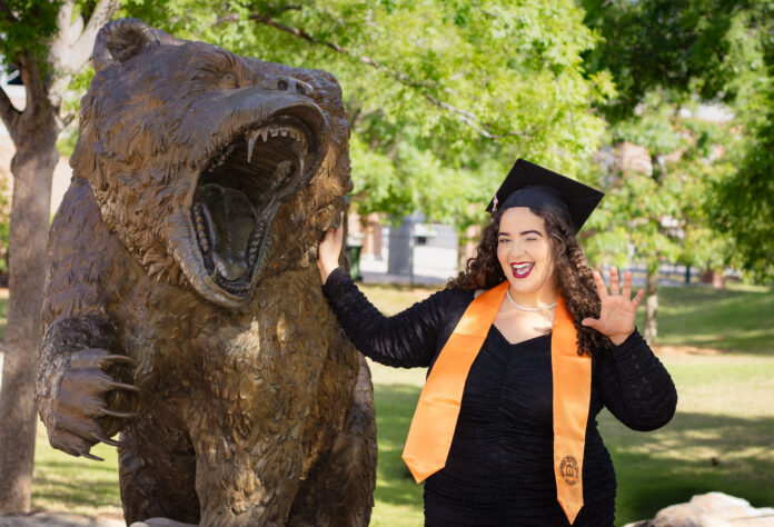 A graduate in a cap and gown poses next to a roaring bear statue outdoors, smiling and holding up one hand.
