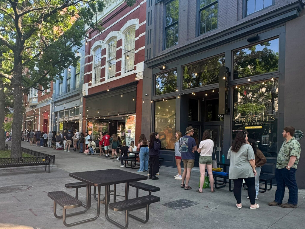 People stand in a long line outside storefronts on a city street near trees and outdoor seating on a sunny day.