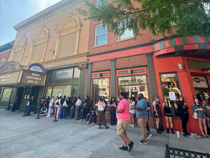 A group of people waits in line outside a theater and nearby restaurants on a city sidewalk.