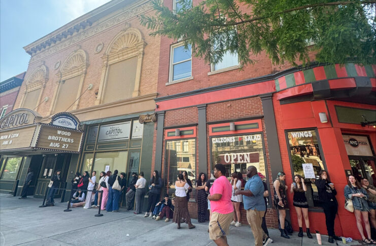A group of people waits in line outside a theater and nearby restaurants on a city sidewalk.