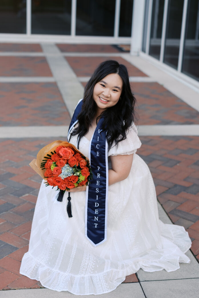 Woman in a white dress kneeling on brick pavement, holding a bouquet, wearing a sash that reads Vice President.