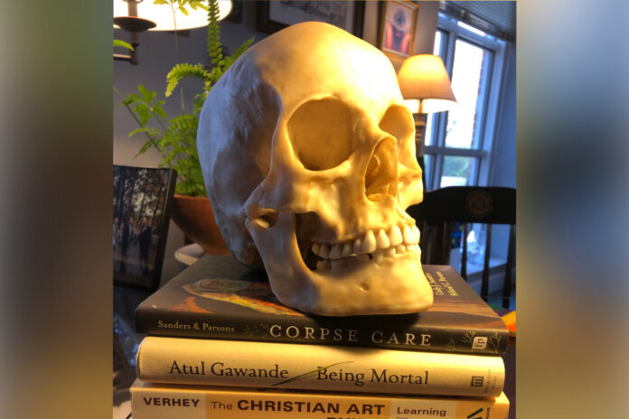 A realistic skull model rests atop three books titled Corpse Care, Being Mortal, and The Christian Art of Dying.
