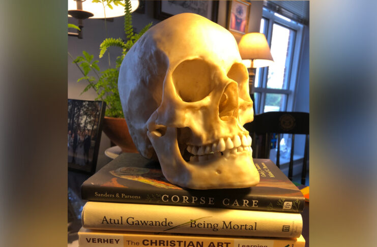 A realistic skull model rests atop three books titled Corpse Care, Being Mortal, and The Christian Art of Dying.
