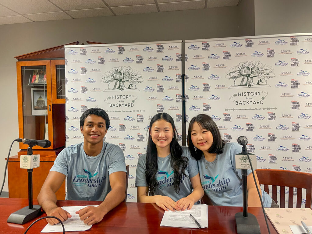 Three students sit at a table with microphones and paperwork, wearing matching Leadership shirts, in front of a branded backdrop.