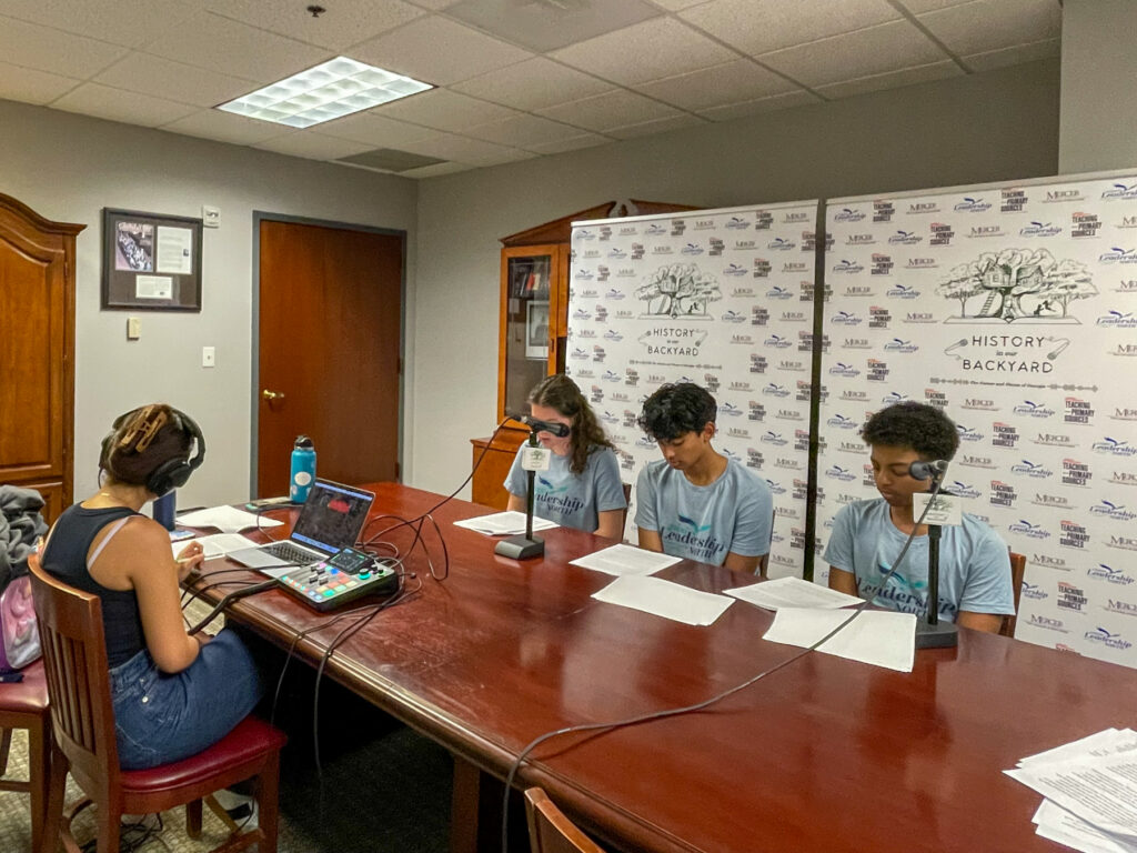 Four young people sit at a table with microphones and papers, recording a podcast in an office setting.
