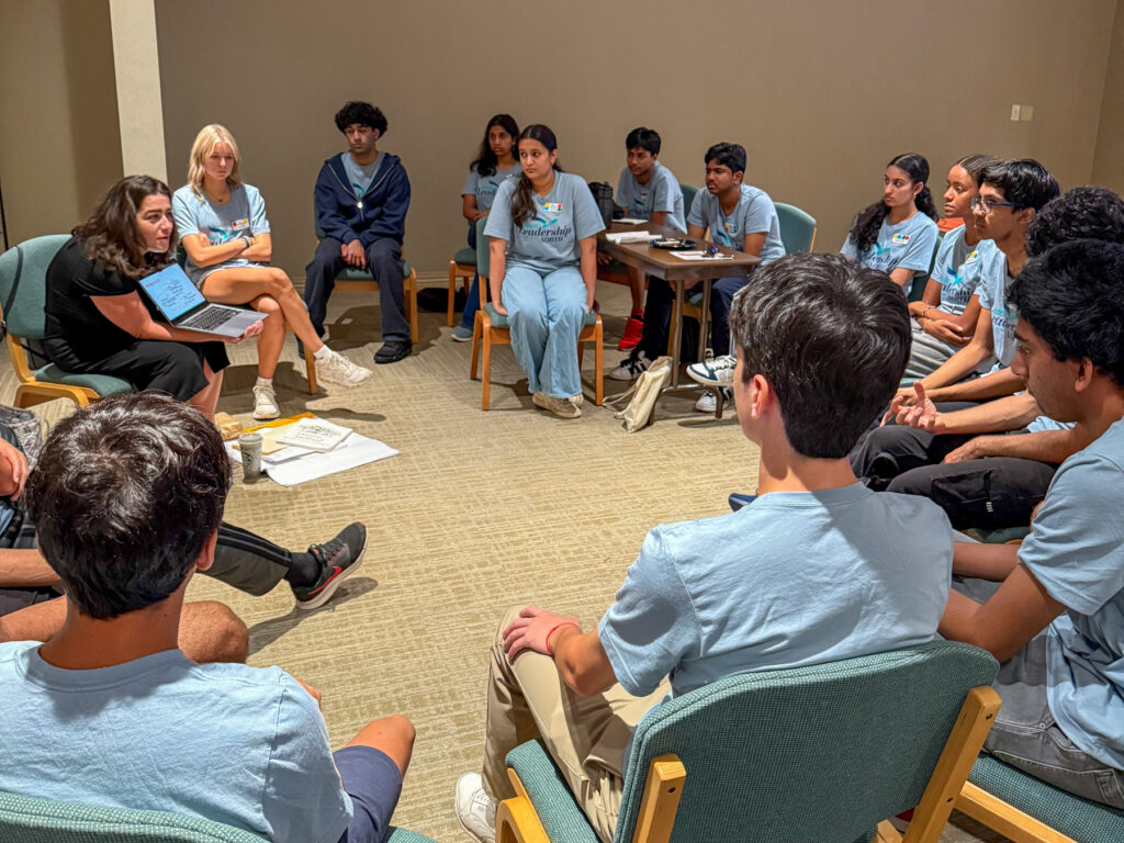 A group of students sit in a circle in a classroom, listening to an instructor holding a laptop and speaking.