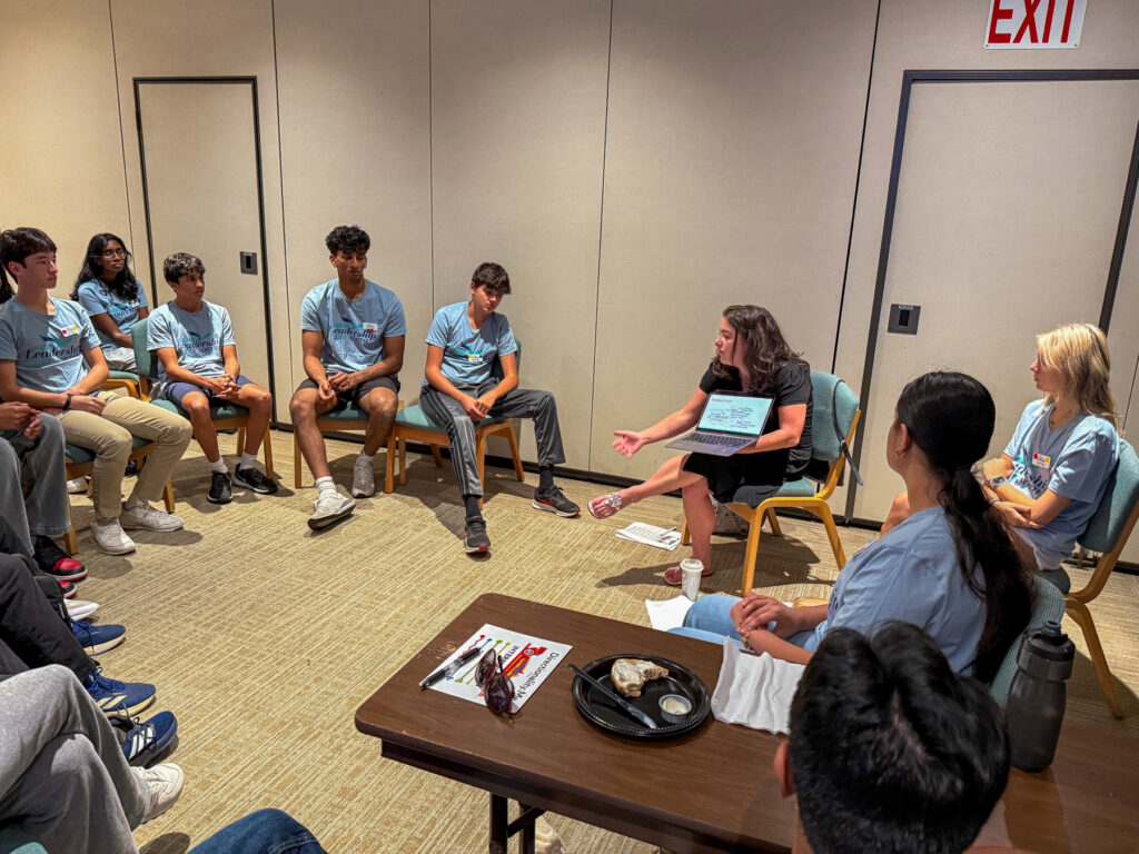 A group of people in blue shirts sit in a circle having a discussion in a conference room.