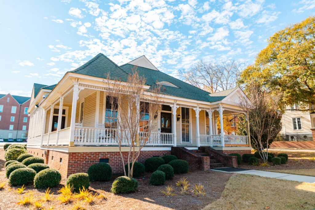 A large, single-story house with a wraparound porch, brick foundation, and green roof on a sunny day.