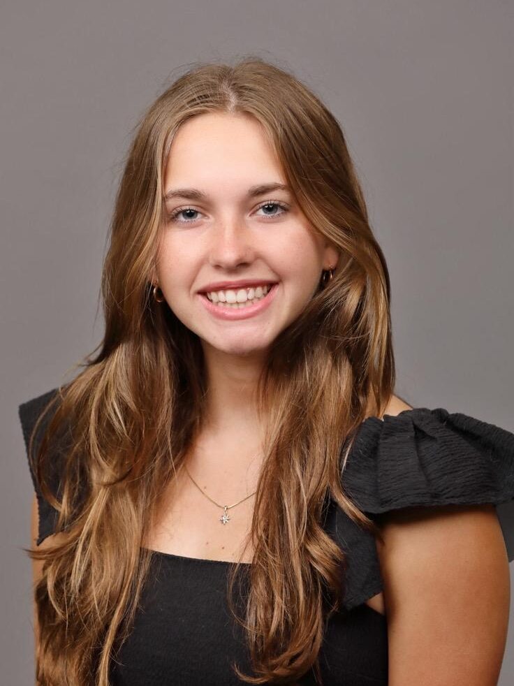 Young woman with long brown hair and a black top smiles at the camera against a plain gray background.