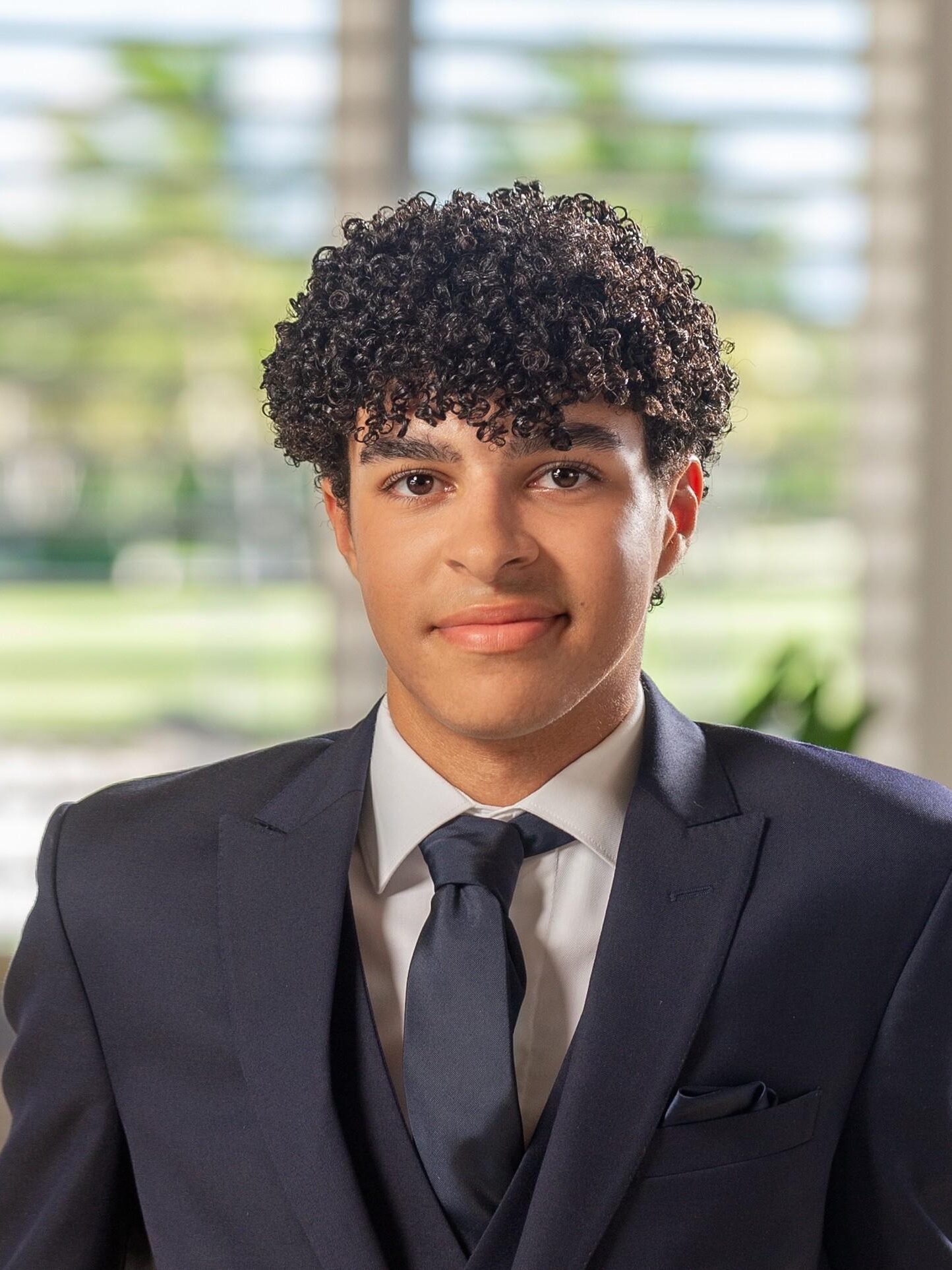 A young man with curly hair in a navy suit and tie stands indoors in front of a window with blinds.