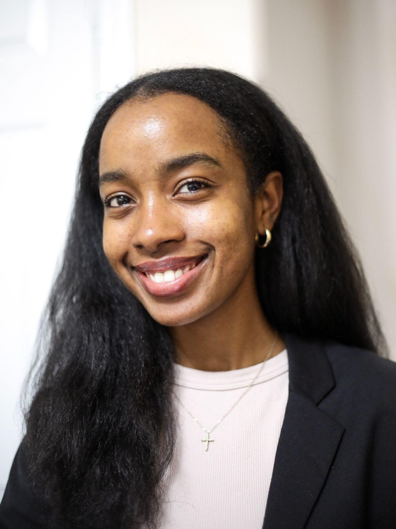 A woman with long dark hair smiles at the camera, wearing a black blazer and a gold cross necklace.