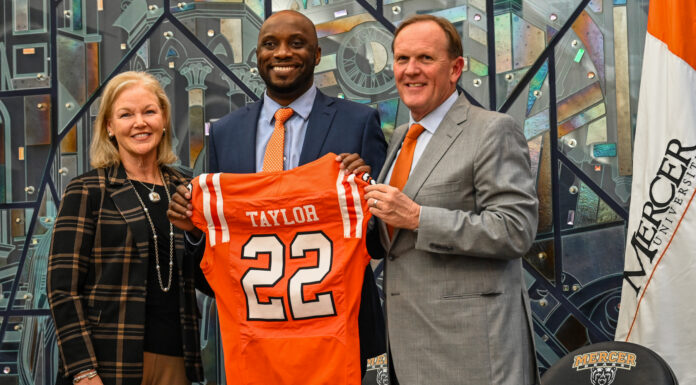 New head coach readies Mercer football for historic run President Dr. Penny L. Elkins, Joel Taylor and Jim Cole pose indoors as Joel Taylor holds a Mercer University football jersey labeled Taylor 22.