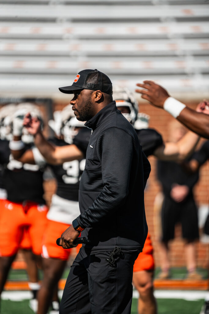Joel Taylor observes players practicing on a field, with athletes in the background.