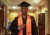Graduate Spotlight: Michael Bloom A graduate in cap and gown with an orange stole holds a conductors baton, standing indoors and smiling at the camera.