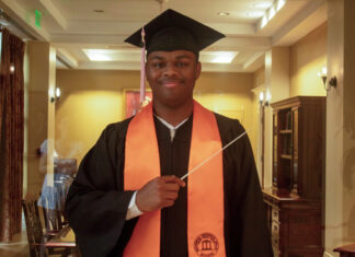 Graduate Spotlight: Michael Bloom A graduate in cap and gown with an orange stole holds a conductors baton, standing indoors and smiling at the camera.