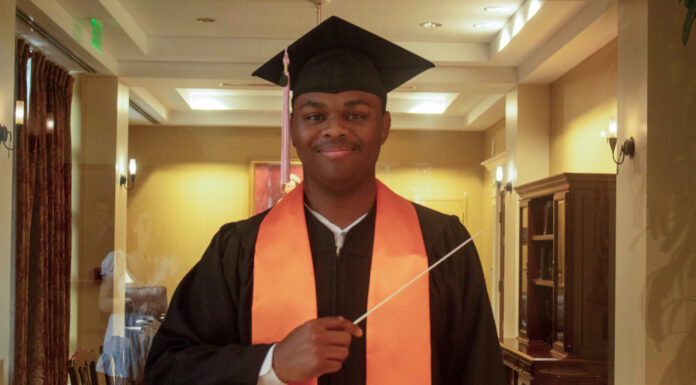 Graduate Spotlight: Michael Bloom A graduate in cap and gown with an orange stole holds a conductors baton, standing indoors and smiling at the camera.
