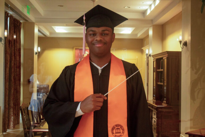 A graduate in cap and gown with an orange stole holds a conductors baton, standing indoors and smiling at the camera.