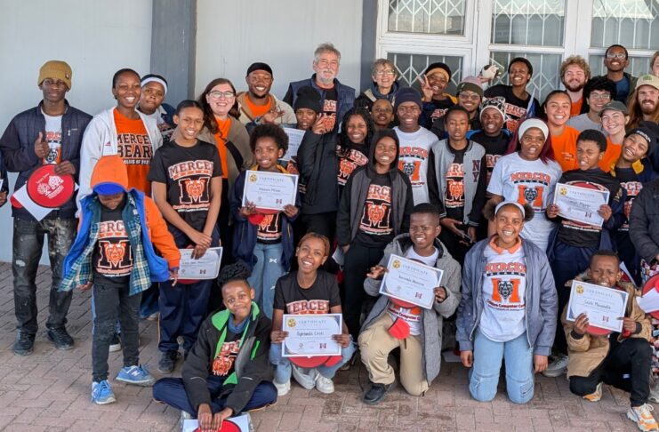 A large group of children and adults pose together outside a building, holding certificates and wearing matching shirts.
