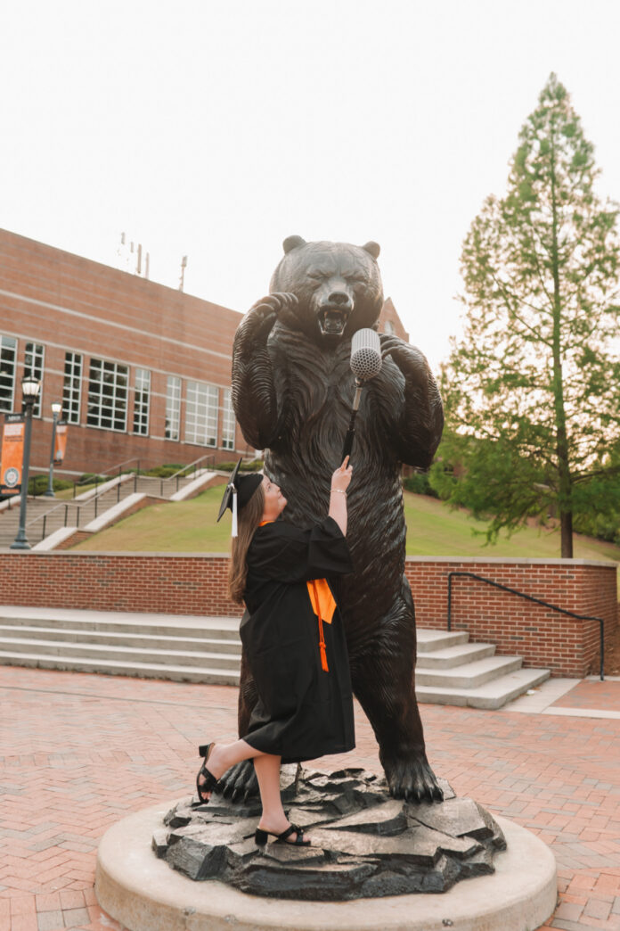 Pariss Nicholson in cap and gown poses playfully with a microphone in front of the bear statue at Mercer.