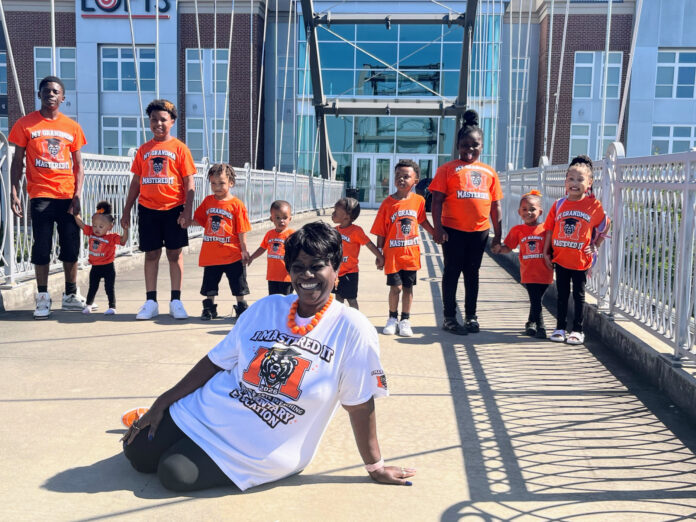 A group of children and one adult pose on a bridge, all wearing orange T-shirts except the seated woman in white.