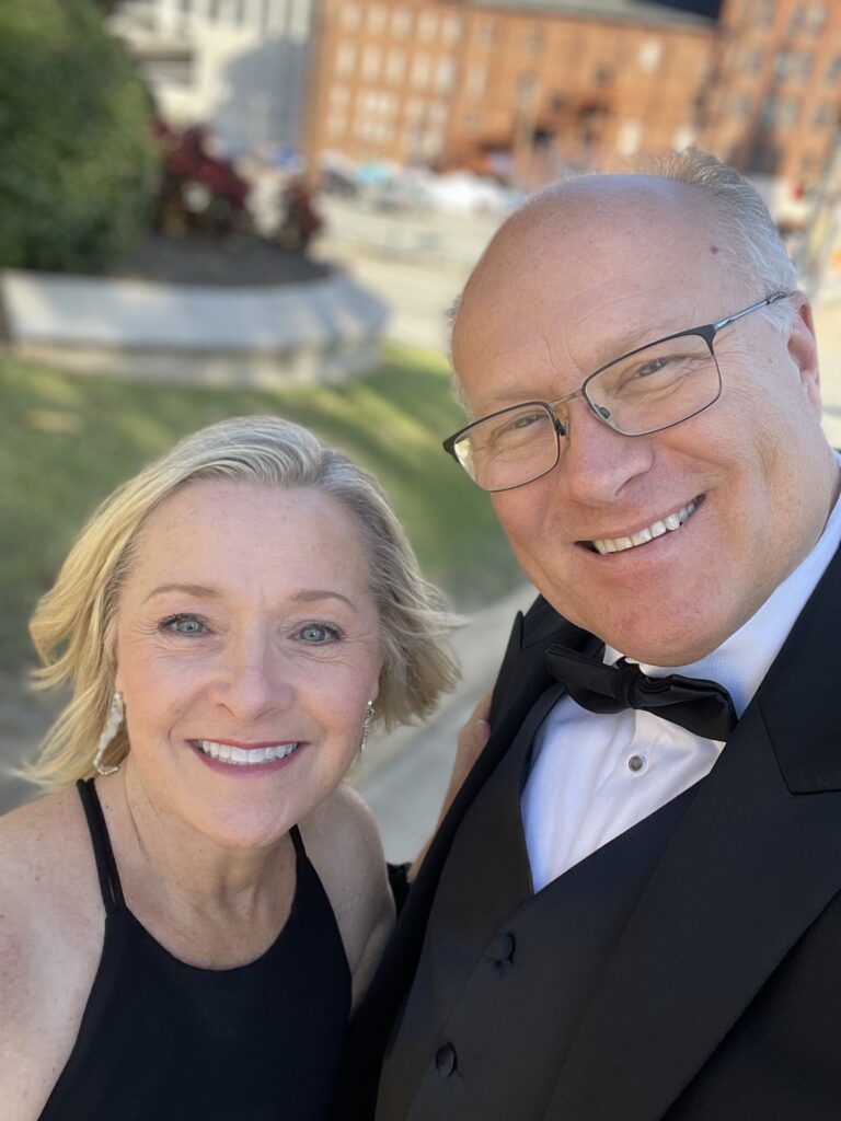 A smiling man in a tuxedo and a woman in a black dress take a selfie outdoors on a sunny day.