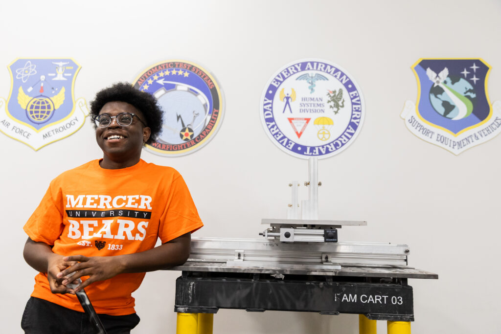 A person in a Mercer University Bears shirt stands by lab equipment, with Air Force-related logos on the wall behind them.